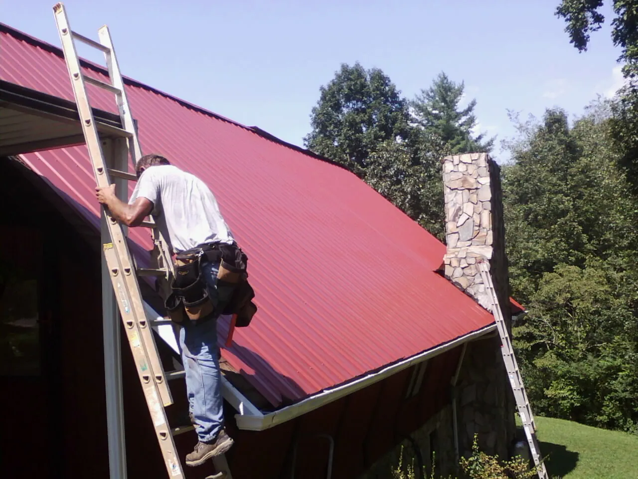 Roofer on a ladder beside a red metal roof and stone chimney — Soffit & Fascia Repair by Roofing Brothers in Waynesburg